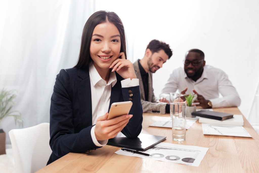 beautiful asian businesswoman in office joyfully looking in camera with cellphone in hands